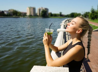 Şehir parkında limonata içen genç bir kadın. Rahatlayan kız nane yapraklı alkolsüz antioksidan içeceği içiyor. Güneşli bir yaz günü. Şehir göletinin seti. Koşudan sonra sportif kadın