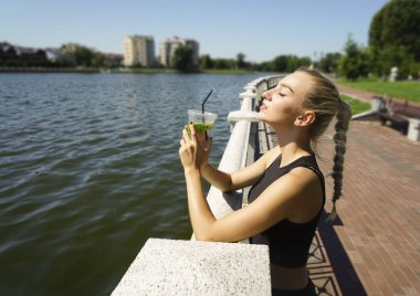 Şehir parkında limonata içen genç bir kadın. Rahatlayan kız nane yapraklı alkolsüz antioksidan içeceği içiyor. Güneşli bir yaz günü. Şehir göletinin seti. Koşudan sonra sportif kadın