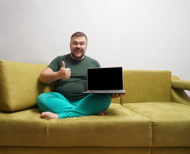 Orta yaşlı mutlu bir adam siyah ekranlı bir dizüstü bilgisayarın arkasında ve koltukta otururken kameraya baş parmağını gösteriyor..