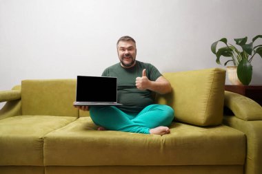 Orta yaşlı mutlu bir adam siyah ekranlı bir dizüstü bilgisayarın arkasında ve koltukta otururken kameraya baş parmağını gösteriyor..