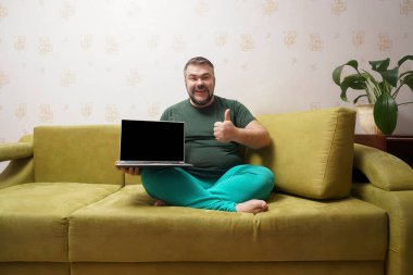 Orta yaşlı mutlu bir adam siyah ekranlı bir dizüstü bilgisayarın arkasında ve koltukta otururken kameraya baş parmağını gösteriyor..