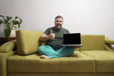 Orta yaşlı mutlu bir adam siyah ekranlı bir dizüstü bilgisayarın arkasında ve koltukta otururken kameraya baş parmağını gösteriyor..