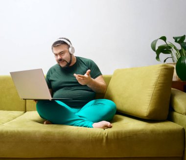 Ciddi sakallı, dizüstü bilgisayarlı, kablosuz kulaklıklı ve günlük kıyafetleriyle koltukta tek başına oturmuş en sevdiği müziği dinleyen bir erkek. 