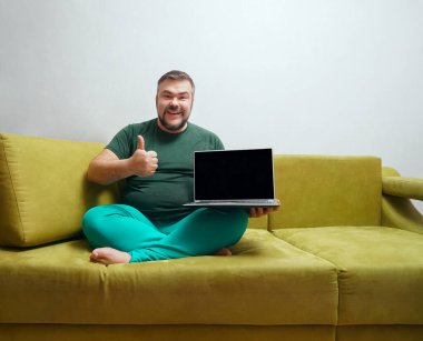 Orta yaşlı mutlu bir adam siyah ekranlı bir dizüstü bilgisayarın arkasında ve koltukta otururken kameraya baş parmağını gösteriyor..