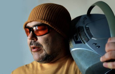 Pozitif ruh hali olan genç bir adamın fotoğrafı beyaz ya da gri arka planda izole edilmiş omuzlarında teyp taşıyor. Sarı şapkalı, güneş gözlüklü ve müzik dinleyen bir adam..