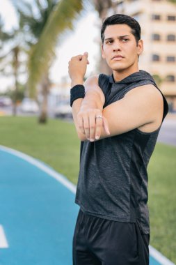 Latin kökenli genç bir adam parkta egzersiz yapmadan önce kollarını açıyor.