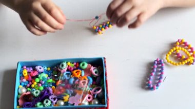 Küçük çocuk elleri çok renkli boncuklardan bir bilezik yapar. Üzerinde çok renkli boncuklar olan beyaz bir kutu.