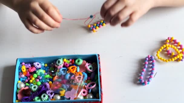 Kleine kinderhandjes maken een armband van multi-gekleurde kralen. Een doos met multi-gekleurde kralen op een witte tafel — Stockvideo