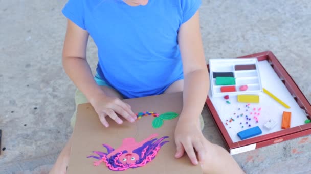 Он-лайн тренинг в творчестве, ребенок лепит на улице. Ребенок делает рисунок из пластилина, на котором изображена разноцветная русалка. — стоковое видео