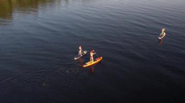 Gün batımında Stand Up Paddle 'da veya nehirde SUP sörf yapan insanların hava görüntüsü. Yaz tatilinde deniz kenarında su sporu yapan kadın ve erkeğin silueti. Sağlıklı yaşam tarzı Aktif turizm