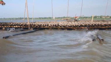 Kırsal kesimde (rüzgar ve akım koşulları) bir nehir boyunca inşa edilmiş bir bambu köprüsü üzerinde su seviyesi yakın çekim görüntüsü; ağır yük taşıyan bir at arabası köprüden geçiyor