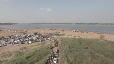 Yerel geçiş ücreti bambu köprüsünde bekleyen uzun araç kuyruğunun hava aracı görüntüsü. Kalabalık yüzmek için toplandı. Kum plajına yayılmış ahşap bungalovlar var.