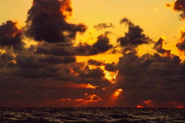 Dalgalanan denizin yüzeyi ufka ulaşıyor. Gökyüzünde, yükselen güneşin resimsel ışınlarının içinden geçtiği karanlık bulutlar var..