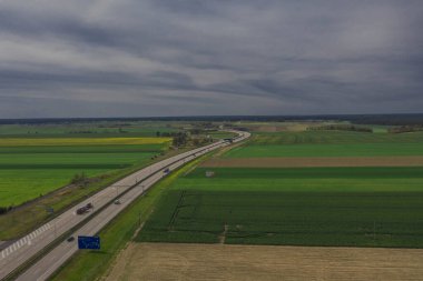 Güneşli bir gün. Gökyüzü biraz bulutlu, ovalar ekilebilir tarlalar ve çayırlarla kaplı. Ovada iki şeritli beton bir yol var. Üzerinde giden arabaları görebilirsin..
