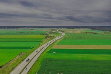Güneşli bir gün. Gökyüzü biraz bulutlu, ovalar ekilebilir tarlalar ve çayırlarla kaplı. Ovada iki şeritli beton bir yol var. Üzerinde giden arabaları görebilirsin..