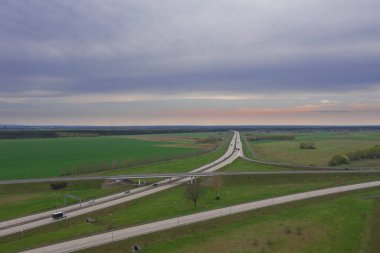 Güneşli bir gün. Gökyüzü biraz bulutlu, ovalar ekilebilir tarlalar ve çayırlarla kaplı. Ovada iki şeritli beton bir yol var. Üzerinde giden arabaları görebilirsin..