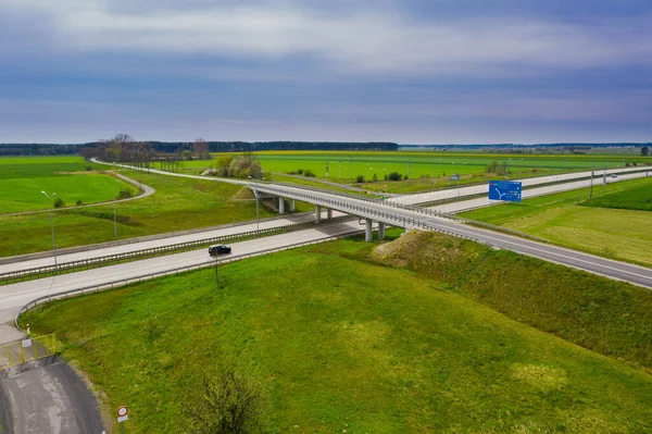Güneşli bir gün. Gökyüzü biraz bulutlu, ovalar ekilebilir tarlalar ve çayırlarla kaplı. Ovada iki şeritli beton bir yol var. Üzerinde giden arabaları görebilirsin..