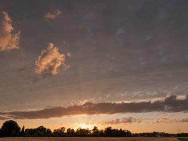 Slightly cloudy evening sky over the plain. The sun is low over the horizon, giving a red light. Streaks of light break through the clouds.