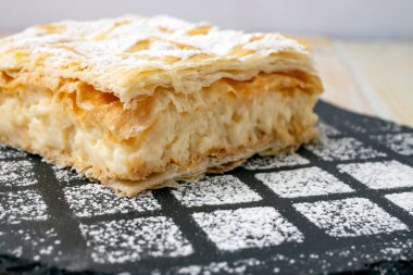 Close up of a cremeschnitte or cremsnit cake on a black stone tile texture with sprinkled sugar powder 
