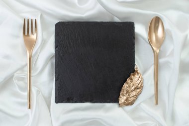 Restoran sunumu ya da menüsü için şık bir broşür. Altın çatalı, kaşığı ve gümüş saten arka planda bir yaprağı olan kare siyah taş döşeme.