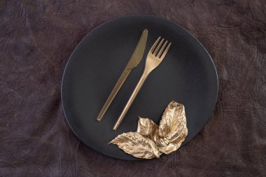 Altın çatal bıçak takımı ve yapraklar siyah seramik tabakta, kahverengi deri arka planda.
