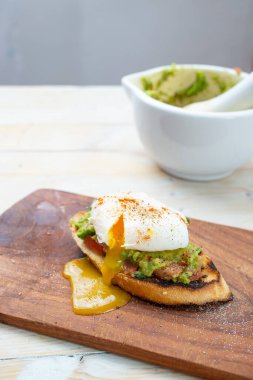 Bir dilim tost ekmeğinde haşlanmış yumurta, guacamole ve yumurta sarısı damlayan ahşap doğrama tahtasının üzerinde. Arkaplanda beyaz harç ve tokmak
