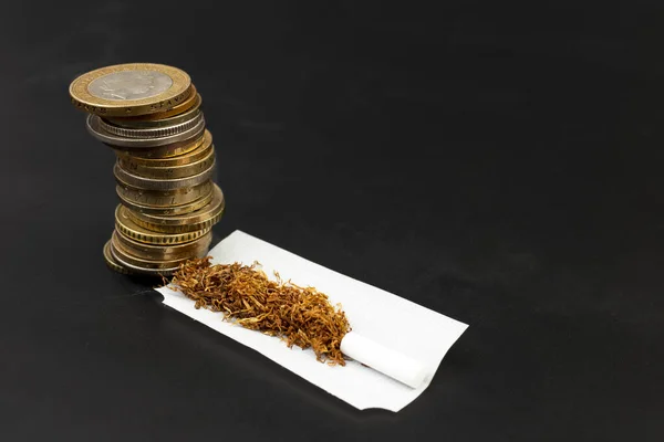 Rolling paper and tobacco next to a stack of coins isolated on black. Money going up in smoke metaphor