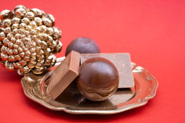 Koyu çikolata topları ve kareler altın tepside, kırmızı arka planda bir kozalağın yanında. 