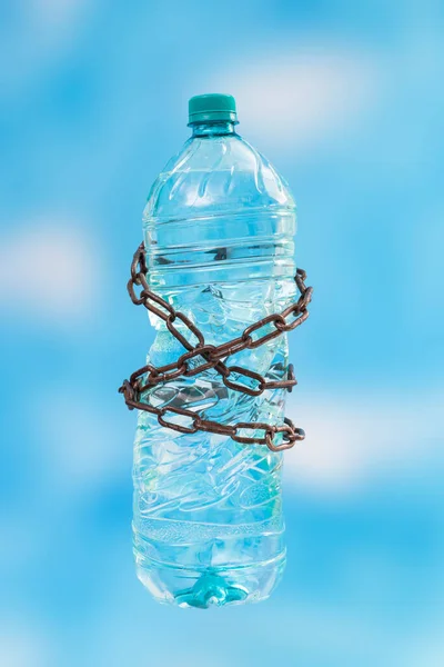 Plastic water bottle tied with rusty chains on blue skyes background. Water crisis metaphor.