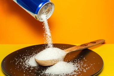 Pouring crystal sugar from a blue can to a wooden spoon on a black plate and orange background.  Motion blur on sugar texture. Sugar danger concept