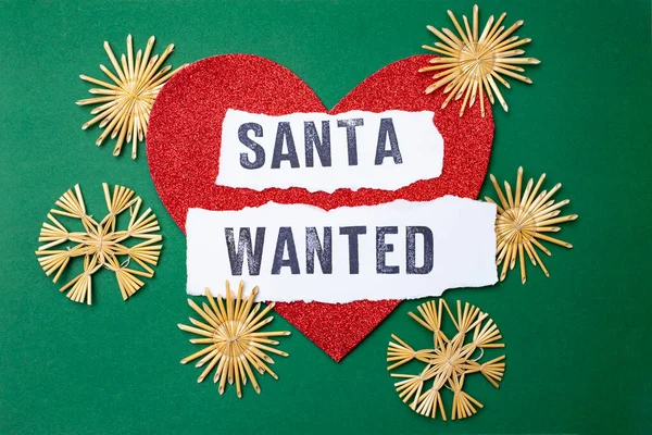 Santa wanted written on white paper strips, on a red glitter heart and green background. Santa for hire sign