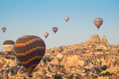 GOREME, TURKEY - 5 AĞUSTOS 2021: Kapadokya 'daki Goreme şehrinin üzerinde sabah ışığında Uchisar Kalesi' nin uzak arkaplanda uçuşan renkli sıcak hava balonları