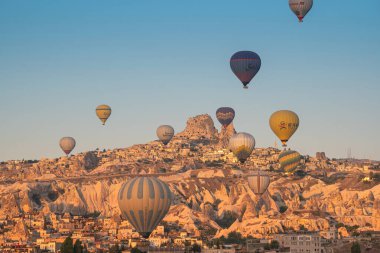 GOREME, TURKEY - 5 AĞUSTOS 2021: Kapadokya 'daki Goreme şehrinin üzerinde sabah ışığında Uchisar Kalesi' nin uzak arkaplanda uçuşan renkli sıcak hava balonları