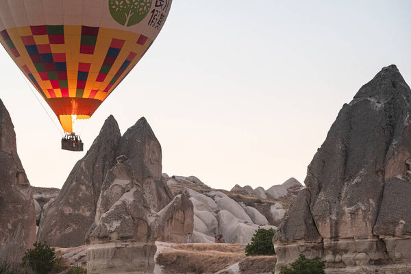 GOREME, TURKEY - AUGUST 5, 2021: Hot air balloon with tourists firing up its burner flame and flying low next to fairy chimneys cave houses in Cappadocia valley in the morning