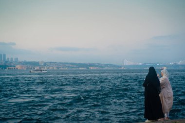 İSTANBUL, TURKEY - 17 Temmuz 2021: Türbanlı iki genç Müslüman kadın yolcu, gün batımında İstanbul 'un arka planında İstanbul kentiyle birlikte İstanbul kıyılarında su kenarında fotoğraf çekiyor