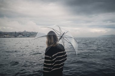Sarı saçlı, siyah kazaklı, şeffaf bir şemsiye tutan ve yağmurlu bir günde denizi izleyen genç beyaz kadının dikiz görüntüsü.