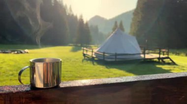 closeup front view of a steaming metallic cup with warm coffee or tea in the morning with glamping camping ground in the background