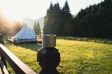 closeup front view of metallic coffee cup in the morning with glamping tent camping ground in the background in a green mountain meadow surrounded by fir tree forest