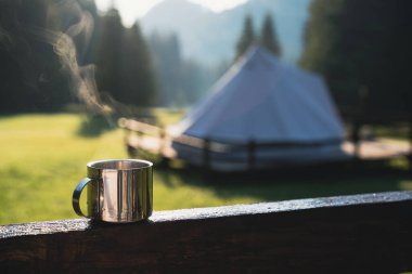 closeup front view of a steaming metallic cup with hot coffee or tea in the morning with glamping camping ground in the background