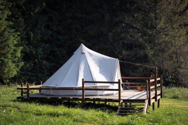 side view of secluded glamping camping tent with white cotton fabric on wooden deck in green meadow surrounded by fir tree forest
