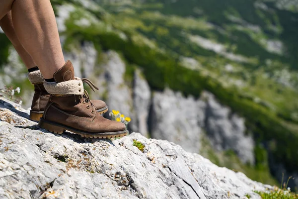 side view closeup detail of Caucasian woman legs with light colored shorts and hiking brown leather boots taking a break on mountain rock