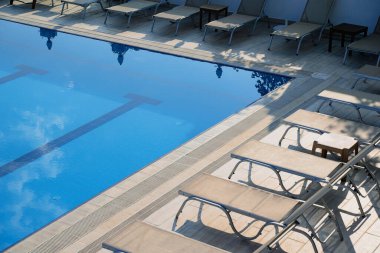 Açık havuz kenarındaki güneşli sandalyelerin yakın plan görüntüsü ve mavi suya yansıyan bulutlu gökyüzü. 