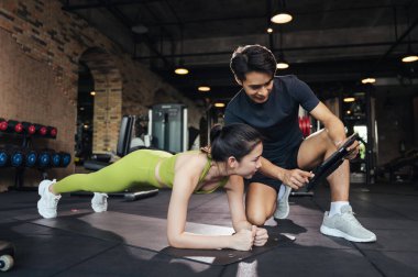 Dijital tablete bakan kişisel antrenör spor salonundaki genç bayana ilerlemeyi açıklıyor. Asyalı erkek antrenör koçu, genç bir bayanın çok yönlü bina zayıf vücudu için planking egzersizine yardım ediyor..