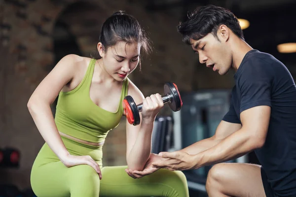 Asian man trainer coach helping sport woman bicep exercise with dumbbell in fitness gym. Healthy lifestyle young female workout training lifting dumbbell with professional trainer.