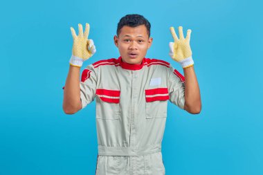 Neşeli genç Asyalı tamirci mavi arka planda izole edilmiş onay sembolü göstererek iyi bir jest yapıyor.
