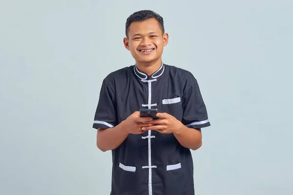 灰色の背景に携帯電話を使用して空手の制服を着て陽気なアジアの若者の肖像画 — ストック写真
