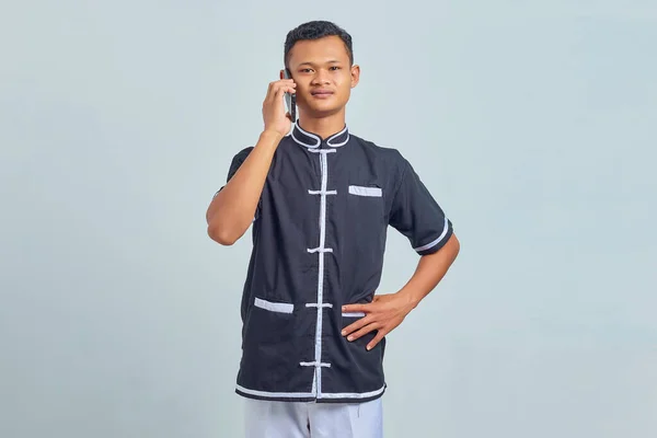 幸せなアジアの若者の肖像空手の制服を着てスマートフォンで話す上で灰色の背景 — ストック写真