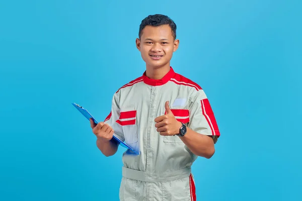 クリップボードを保持し 青色の背景に隔離された親指アップジェスチャーを示す興奮した若いアジアの整備士の肖像 — ストック写真