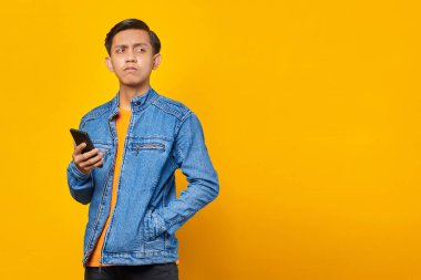 Asyalı adam sarı arka planda cep telefonu tutarken bir şeyler düşünüyor.