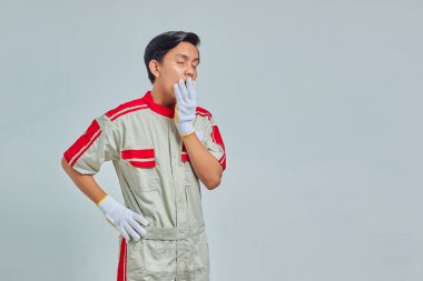 Mekanik üniforma giyen yakışıklı bir adamın portresi uykulu hissediyor ve elleri gri arka planda izole bir şekilde ağzı örtüyor.